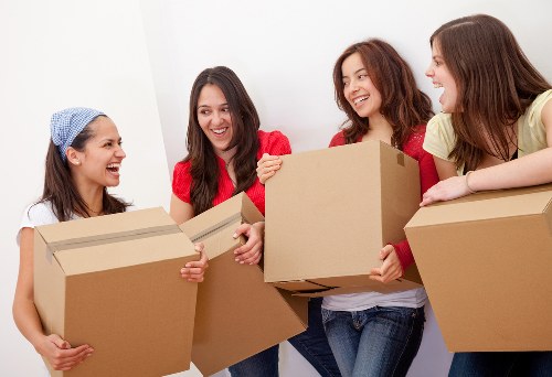 Four young women inside a residential property, engaged in a moving activity, are holding and passing medium-sized cardboard boxes. The woman on the far left, wearing a blue and white patterned headscarf and a white top, is smiling and holding a box close to her chest. The second woman from the left, dressed in a red top, is smiling broadly while extending a box to the right. The third woman, also wearing a red top with long brown hair, is smiling and holding a box at waist height. The woman on the far right, in a light yellow top, is smiling and supporting a box on a flat surface, likely a table or counter. The background features a plain white wall, typical of a home interior, with natural light illuminating the scene, emphasizing the friendly and collaborative atmosphere of a house relocation process. The image visually aligns with house removals services in Earl’s Court, London, showcasing a team or group preparing for a move with cardboard containers, reinforcing themes of household logistics, furniture, and personal belongings being organized for transport.