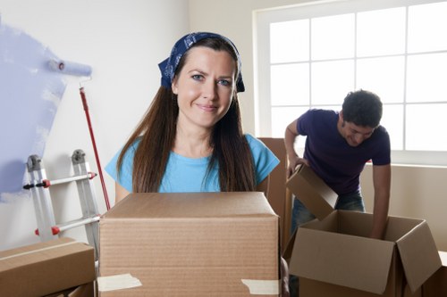 A woman with long brown hair, wearing a blue headscarf and matching blue top, stands in a well-lit room holding a large cardboard box with tape partially applied to its edges. She is smiling gently at the camera. In the background, a man with dark curly hair, dressed in a dark blue T-shirt, is seen leaning forward, handling an open cardboard box as part of a house removal process. The room has a large window allowing natural light to fill the space, and the walls are painted white. To the left, there is a partially visible white wall with a red-handled tool and some shelves or hooks. The scene depicts a professional movers' environment, emphasizing careful packing and handling of household items, consistent with a house removals service in London or nearby areas, with the focus on furniture, boxes, and household belongings typical of a residential move.