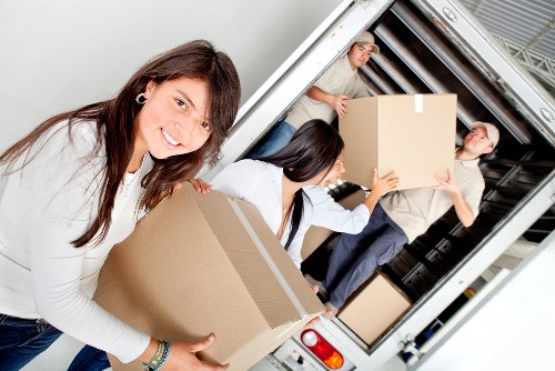 The image shows three professional movers loading cardboard boxes into the back of a white removals van parked outside a residential property, likely in the Earls Court area. In the foreground, a smiling woman with long brown hair, wearing a white shirt, is holding a medium-sized, rectangular cardboard box with a plain brown surface. Behind her, two other movers are visible; one man wearing a light grey shirt with rolled-up sleeves and dark trousers is lifting a large, square cardboard box, while another woman with dark hair in a ponytail, also in a white shirt, is assisting with the packing. The interior of the van has a metallic, ribbed lining and the back door is open to reveal a spacious cargo area suitable for house removals. The outdoor lighting suggests daytime and the scene conveys a professional, organized move typical of house relocation services in London, possibly near the Earl's Court district. The setting emphasizes careful handling of household items, including boxes and possibly other packing materials, supporting the services offered by Earls Court Man with Van for local house removals and relocation tasks.