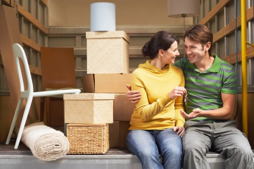 A young couple sitting on the floor of a storage unit surrounded by packed moving boxes and household items, including a woven basket, a rolled-up rug, and a white plastic chair. The woman is wearing a yellow hoodie and blue jeans, while the man is dressed in a green and black striped polo shirt and light grey trousers. They are smiling and engaged in a friendly conversation, with one of them holding a small item. The storage space has wooden shelving on either side, and a neutral background indicating a residential area in the UK, possibly near Earls Court, London. The scene captures a moment of preparing for a house move, with cardboard boxes stacked behind them, some sealed and others open, and soft lighting illuminating the scene, typical of an interior storage environment associated with house removals in the local area.